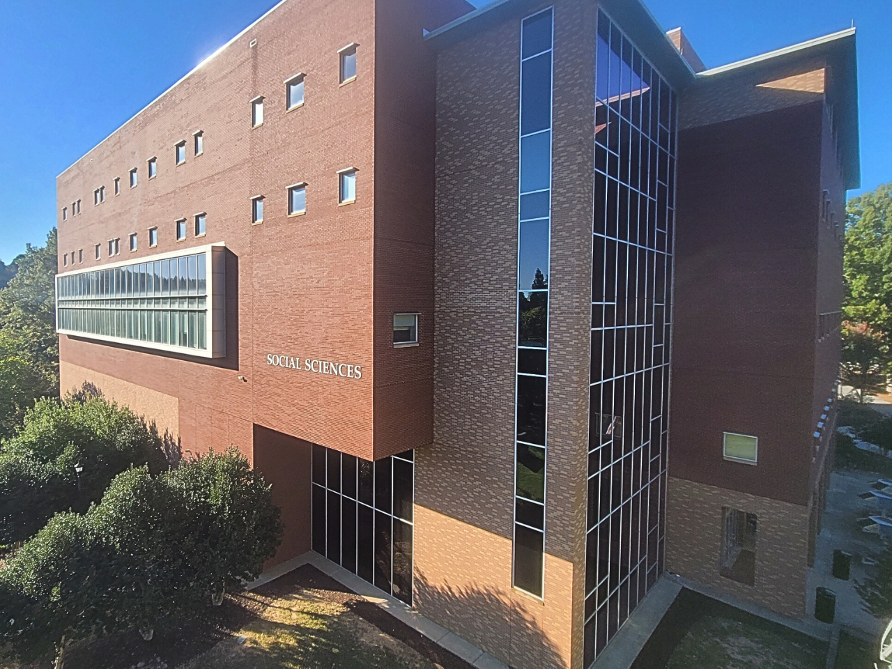 A large brick university building labeled "SOCIAL SCIENCES" features various window styles, including a tall vertical column of glass panes and a long horizontal ribbon window.